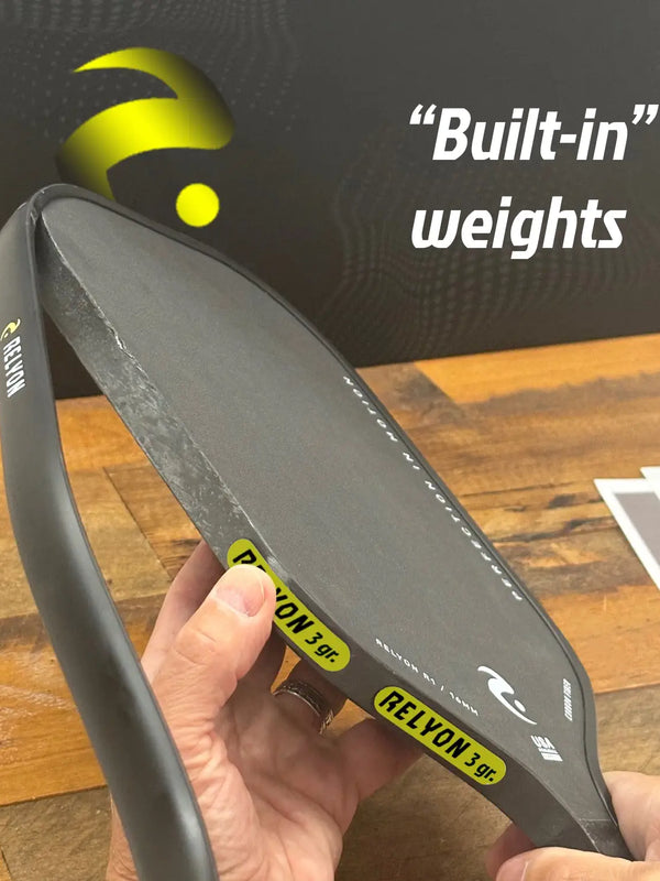 This image shows a person attaching adhesive weight strips to the edge of a Relyon pickleball paddle. The close-up highlights the precision placement of the weights, aimed at adjusting the paddle’s balance and performance. A Relyon-branded box and some papers are partially visible on the wooden surface in the background.