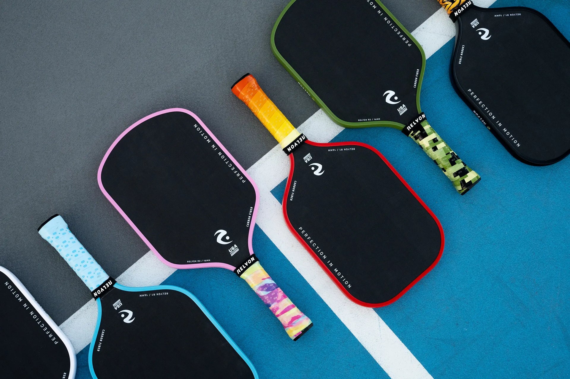 Aerial view of six pickleball paddles arranged in a line on a court, featuring black paddle faces with colorful edges and grips. The designs include pastel blue with dots, pink with a rainbow grip, olive green with a camouflage grip, and red with a gradient orange-yellow grip, all branded
