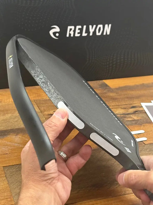 A person adds weight strips to the edge of a Relyon pickleball paddle, customizing its balance and swing weight. The Relyon-branded box sits in the background on a wooden surface.