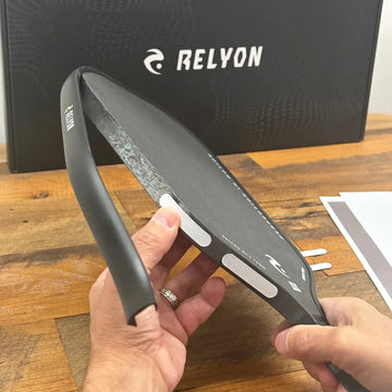 A person adds weight strips to the edge of a Relyon pickleball paddle, customizing its balance and swing weight. The Relyon-branded box sits in the background on a wooden surface.