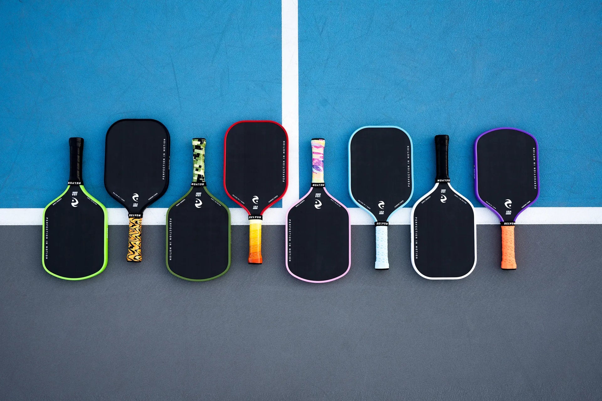  This image showcases a row of eight Relyon pickleball paddles neatly lined up on a court, split between blue and gray surfaces with a white dividing line. The paddles feature black faces, each framed by vibrant edge colors, including green, red, pink, white, and purple. The handles display diverse, eye-catching grips, from solid to patterned designs, reinforcing the brand’s tagline 