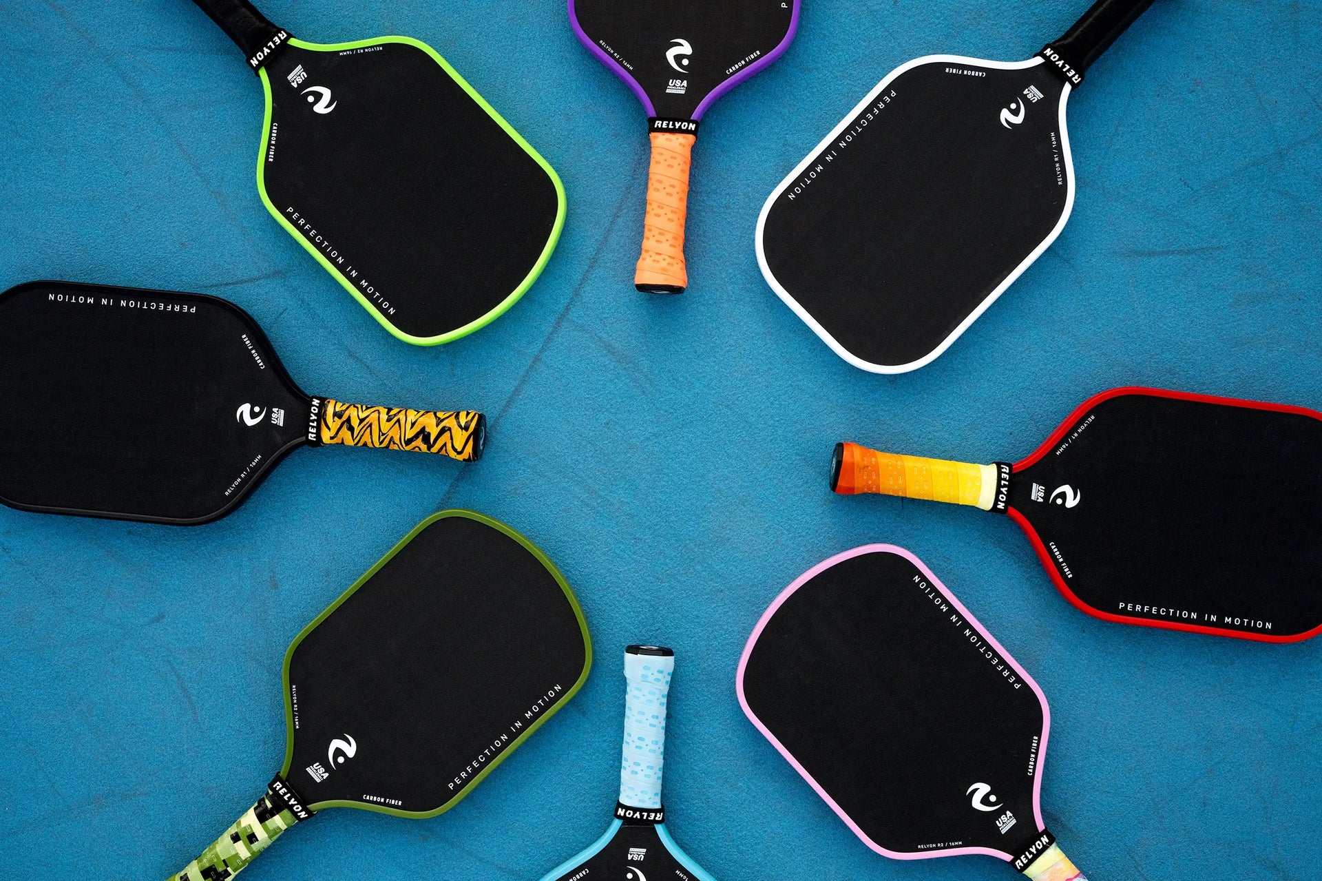  This image shows a variety of pickleball paddles arranged in a circular pattern on a bright blue surface. Each paddle features a black surface with colorful edge guards, including shades of green, purple, white, red, and pink, and vibrantly wrapped handles. The brand name 