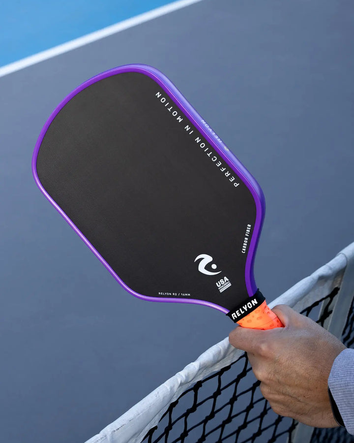 This image features a close-up of a Relyon pickleball paddle with a sleek black face framed by a vibrant purple edge. The paddle, labeled "Relyon R2 / 16MM," highlights its carbon fiber construction and USA Pickleball certification. A hand grips the paddle’s orange handle over the net, emphasizing the slogan “Perfection in Motion” printed along the edge, symbolizing performance and precision on the court.