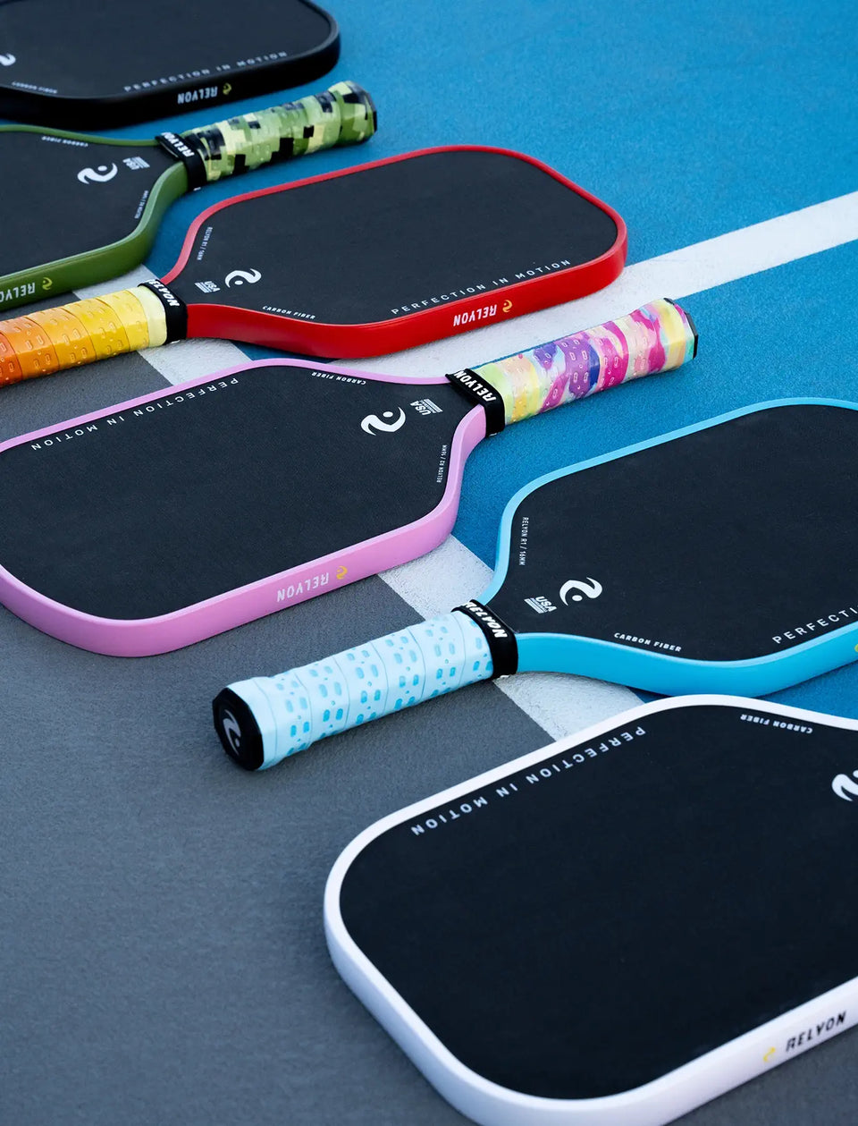  This image displays a collection of Relyon pickleball paddles lined up diagonally along the boundary of a blue and gray court. The paddles showcase sleek black faces with colored edges in green, red, pink, light blue, and white, each paired with unique grip designs, including camouflage patterns and colorful prints. The branding “Perfection in Motion” is visible on each paddle, emphasizing both aesthetics and high-quality performance.