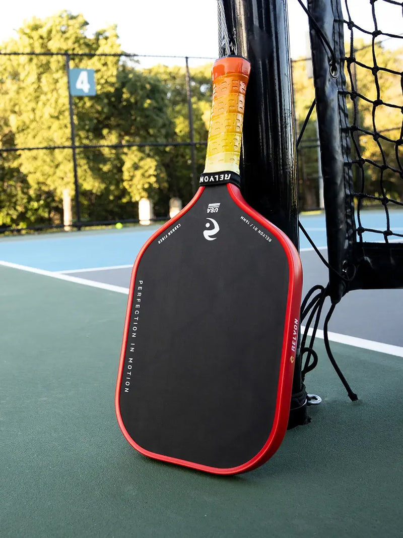 A pickleball paddle with a sleek black face and red trim leans against the net post on an outdoor court. The grip is a gradient of yellow to orange, marked with the brand "RELYON" and the phrase "PERFECTION IN MOTION" printed on the paddle’s edge.