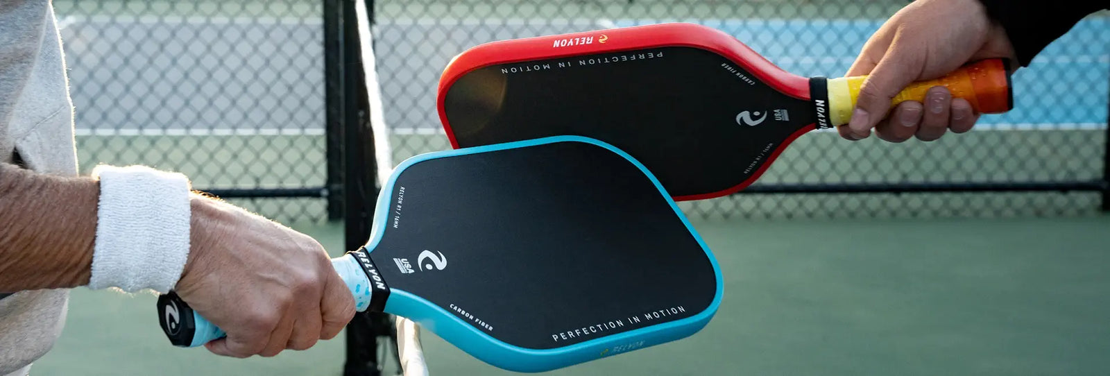 Two players bump their pickleball paddles, one with a red and black paddle and the other with a blue and black paddle. Both paddles feature the "RELYON" branding, the slogan "PERFECTION IN MOTION," and have sleek designs, with the blue paddle's grip matching the paddle's trim and the red paddle's grip transitioning from yellow to orange.