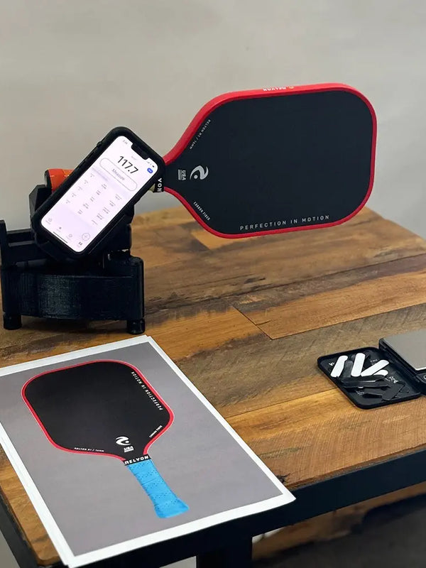 This image shows the swing weight of a Relyon pickleball paddle being measured, with the paddle mounted on a device holding a smartphone displaying a weight of 117.7. On the table, additional tools, including weight strips, a digital scale, and a printed image of the paddle, are neatly arranged, suggesting customization for optimal performance.