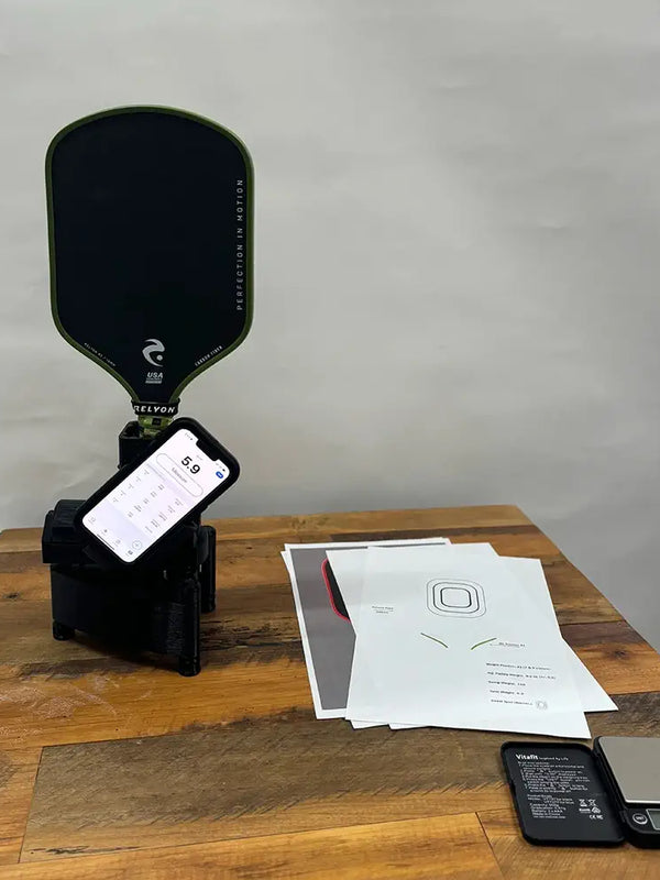 This image shows the twist weight of a Relyon pickleball paddle being measured. The paddle is mounted on a device with a smartphone displaying a reading of 59.9. On the table, a digital scale and printed diagrams are laid out, suggesting careful customization and analysis for optimal paddle performance.