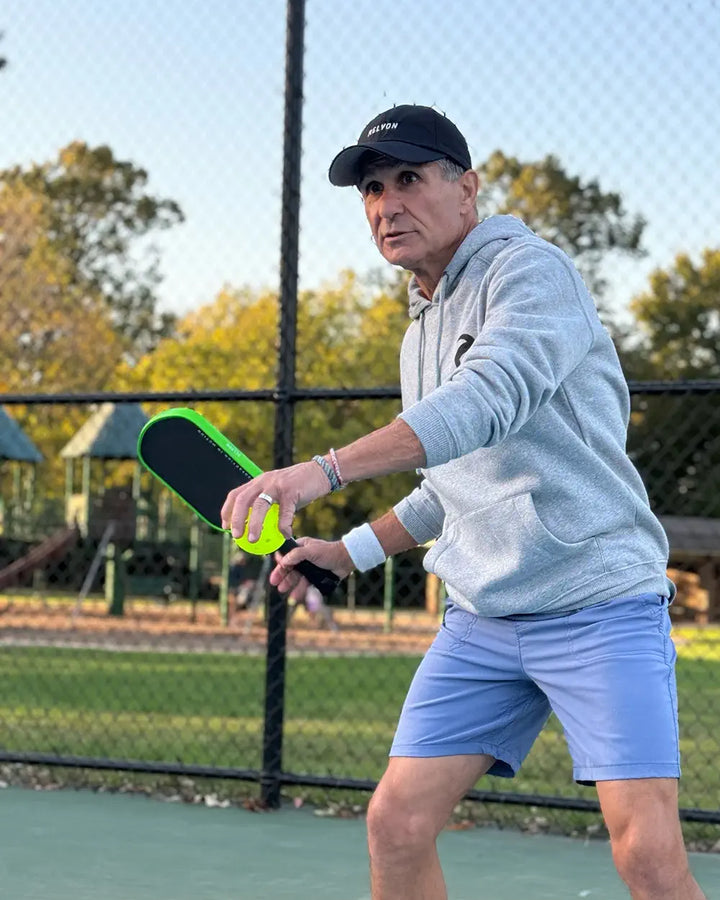 A man in a gray hoodie and blue shorts focuses intently while playing pickleball outdoors. He grips a bright green and black paddle, poised to make his next move on the court. A chain-link fence and park playground are visible in the background, capturing a sunny day at the park.