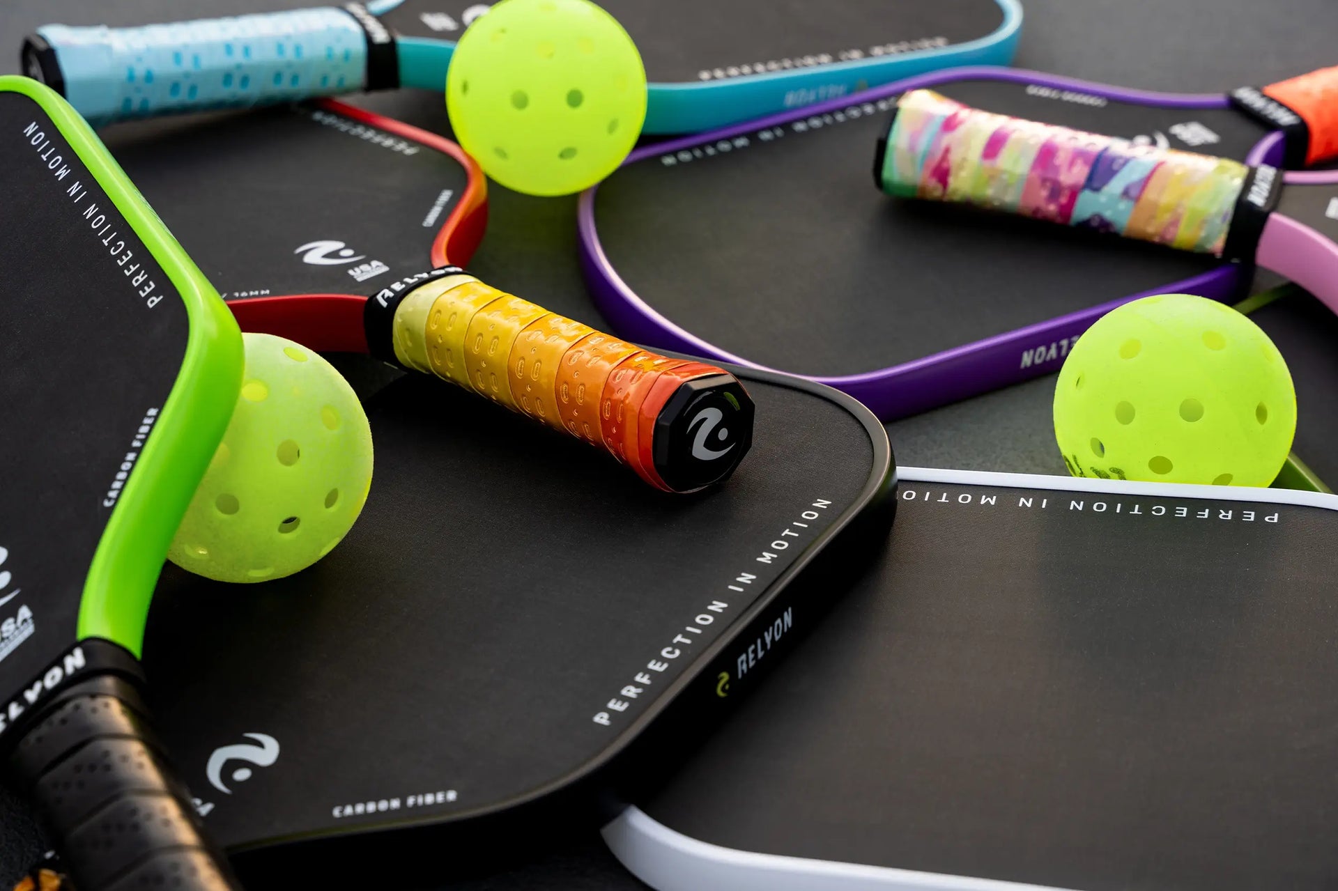 This image features a dynamic arrangement of Relyon pickleball paddles interspersed with bright yellow pickleballs. The paddles, showcasing sleek black faces with colorful edge guards in green, red, purple, and white, highlight the brand’s slogan "Perfection in Motion" along the edge. The variety of colorful, patterned grips adds personality, reinforcing both style and performance, while the scattered pickleballs hint at gameplay excitement.