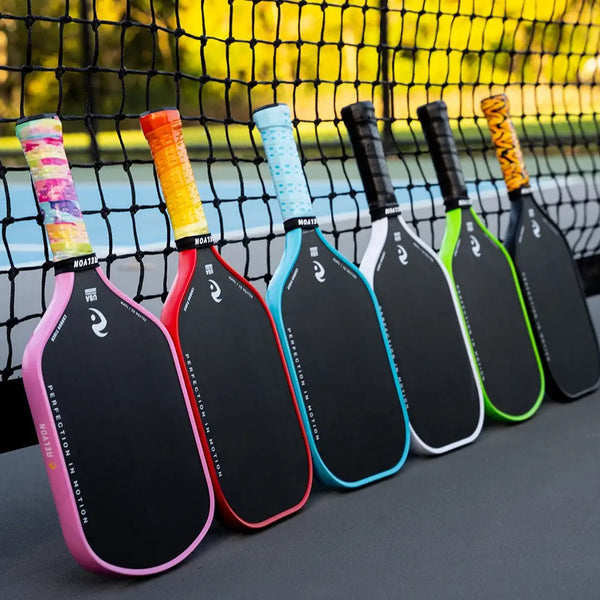 A lineup of six pickleball paddles rests against the net on an outdoor court, each featuring black faces with vibrant, uniquely colored grips and trim. The paddles display colors such as pink, orange, red, blue, black, and green, all branded with "RELYON" and the phrase "PERFECTION IN MOTION" along the paddle faces.