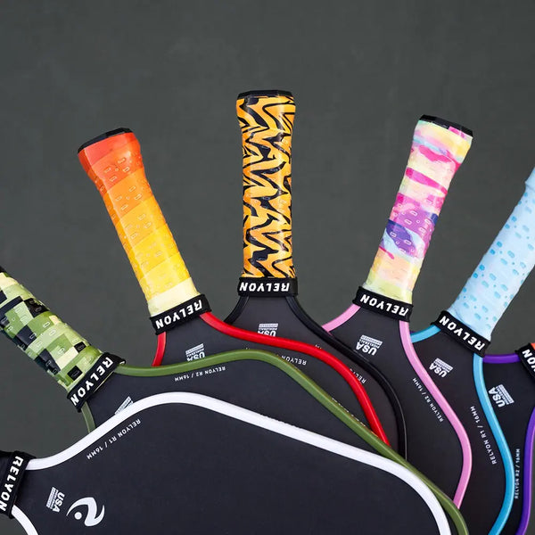 A close-up view of several pickleball paddles highlights their colorful and unique grip tapes. The grips feature various designs, including a gradient yellow-orange, a bold black-and-yellow zigzag, pastel rainbow patterns, and dotted blue. Each paddle is branded "RELYON," emphasizing a modern and vibrant look.