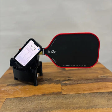 A pickleball paddle with a red trim and black face is mounted on a device used to measure swing weight, with a smartphone displaying a reading of "1177" in large numbers. The paddle is branded "RELYON" with the phrase "PERFECTION IN MOTION," and the setup is displayed on a wooden surface.