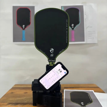 An olive green-trimmed pickleball paddle stands upright on a measurement device, with a smartphone attached showing a reading of "5.9." The paddle, marked "RELYON" and "PERFECTION IN MOTION," is set against a backdrop featuring design mock-ups of other paddles. The setup is displayed on a wooden table, emphasizing a professional testing environment.